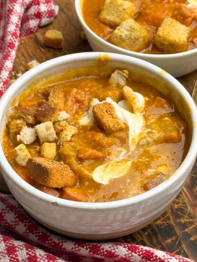 A bowl of thick soup topped with croutons and a swirl of cream sits on a wooden surface near a red and white checkered cloth. Another bowl of soup with croutons is in the background.