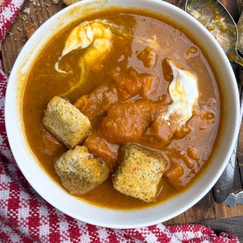 A bowl of thick sweet potato soup, with ham and sweet potatoes, with a dollop of sour cream and several large croutons, on a wooden surface with a red and white checkered cloth and two tarnished spoons nearby.