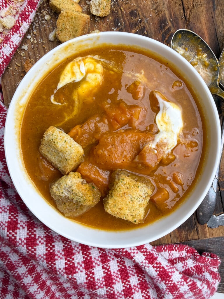 A bowl of thick sweet potato soup, with ham and sweet potatoes, with a dollop of sour cream and several large croutons, on a wooden surface with a red and white checkered cloth and two tarnished spoons nearby.