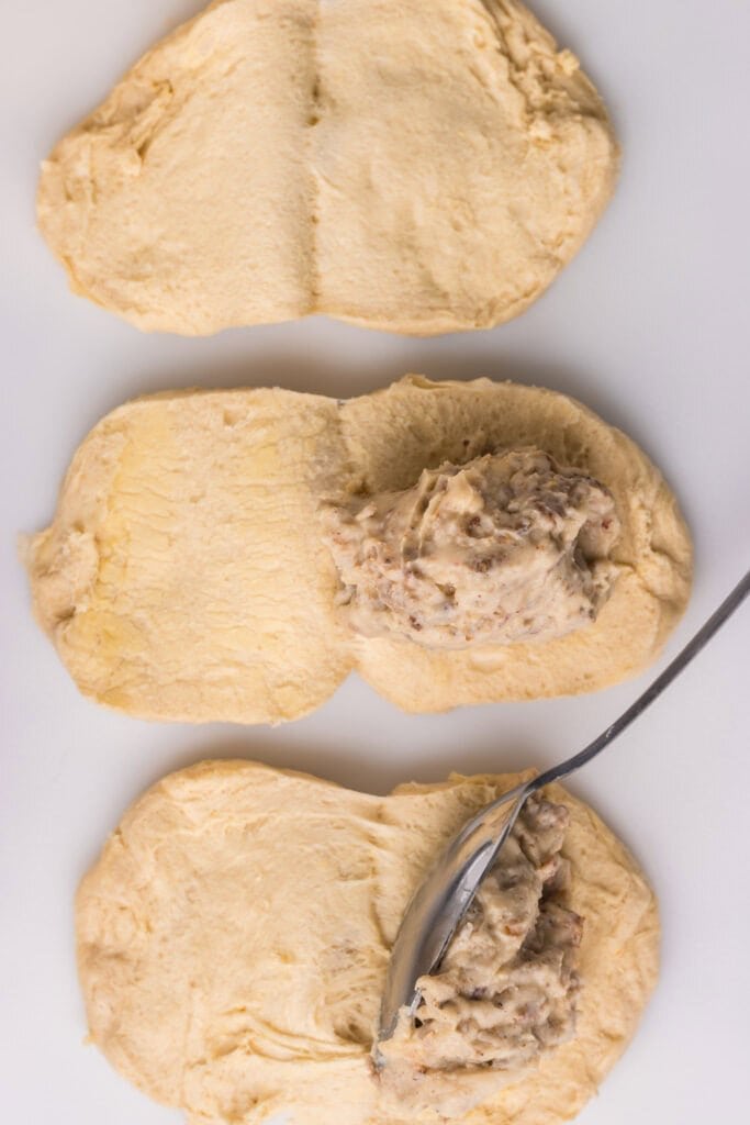 Three pieces of biscuit dough are laid out on a surface, with a spoon spreading a creamy, chunky filling onto the center section of one piece.