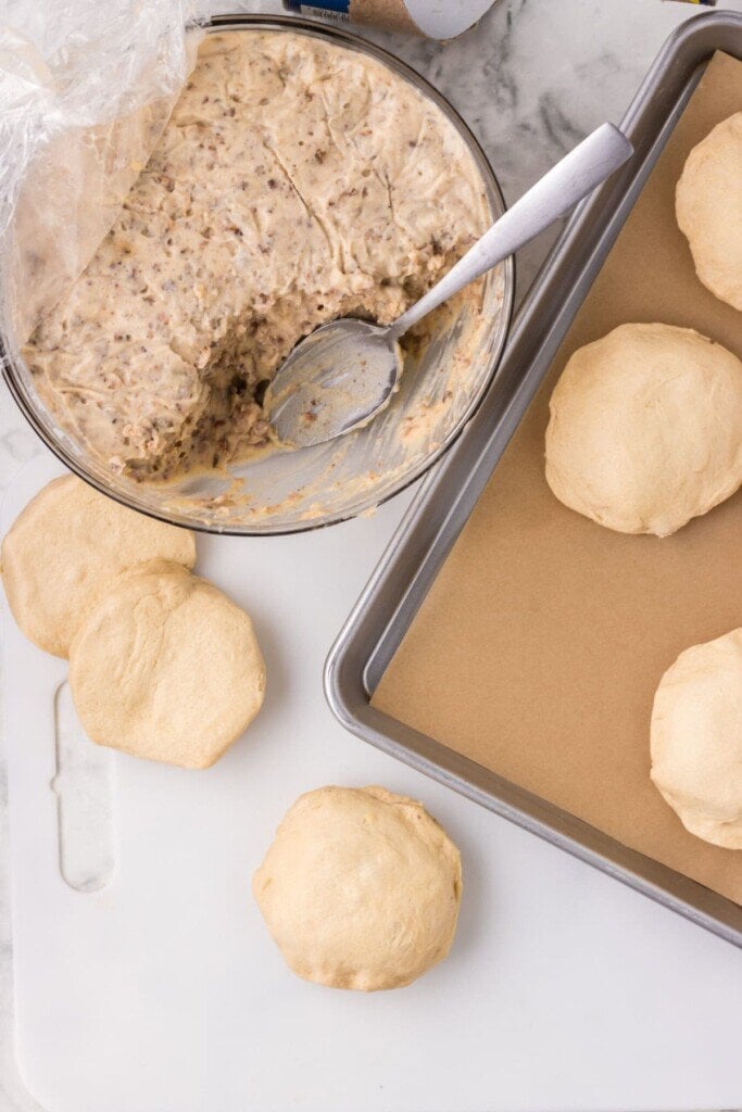 Bowl of creamy sausage gravy with a spoon. Several raw cold sausage biscuit dough balls on a white board, and a baking sheet lined with parchment paper.