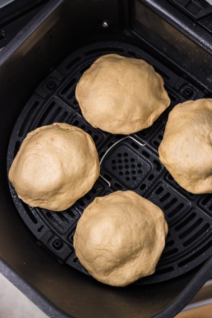 Four pieces of puffy, uncooked dough are arranged inside an air fryer basket, ready to be baked.
