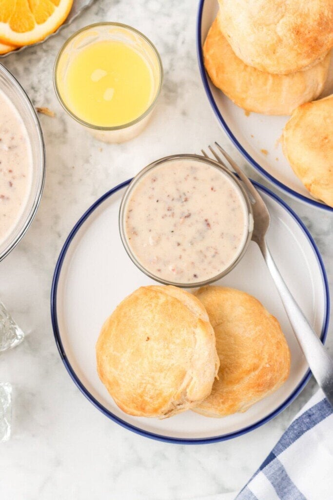 A plate with two Air Fryer Sausage Gravy Stuffed Biscuits, a bowl of creamy gravy, a fork, and a glass of orange juice on a marble table with more biscuits and orange slices nearby. Blue and white napkin in the corner.