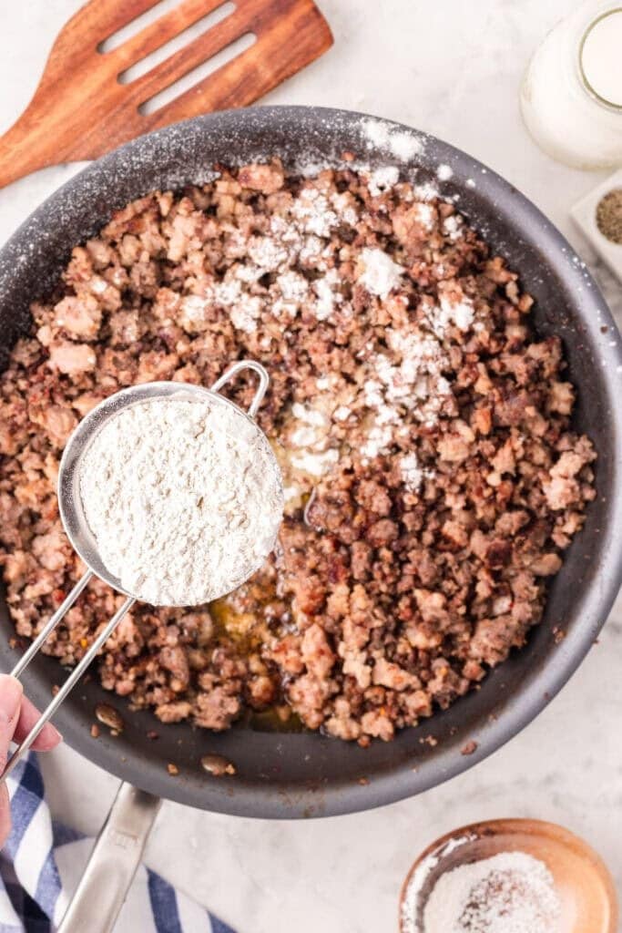 A hand holds a measuring cup of flour over a skillet filled with cooked, crumbled sausage, with some flour already sprinkled on the meat. Kitchen utensils and ingredients are visible nearby.