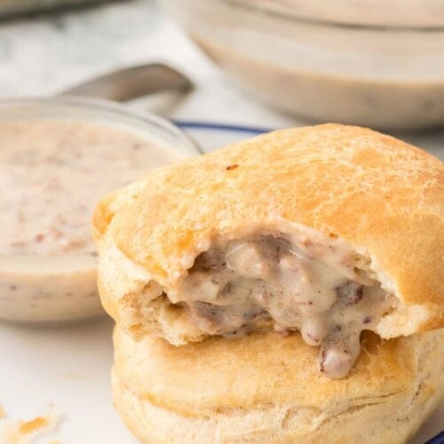 A biscuit with creamy sausage gravy filling, sitting on a white plate. The top biscuit has a bite taken out, showing the gravy inside. A bowl of extra gravy is in the background.