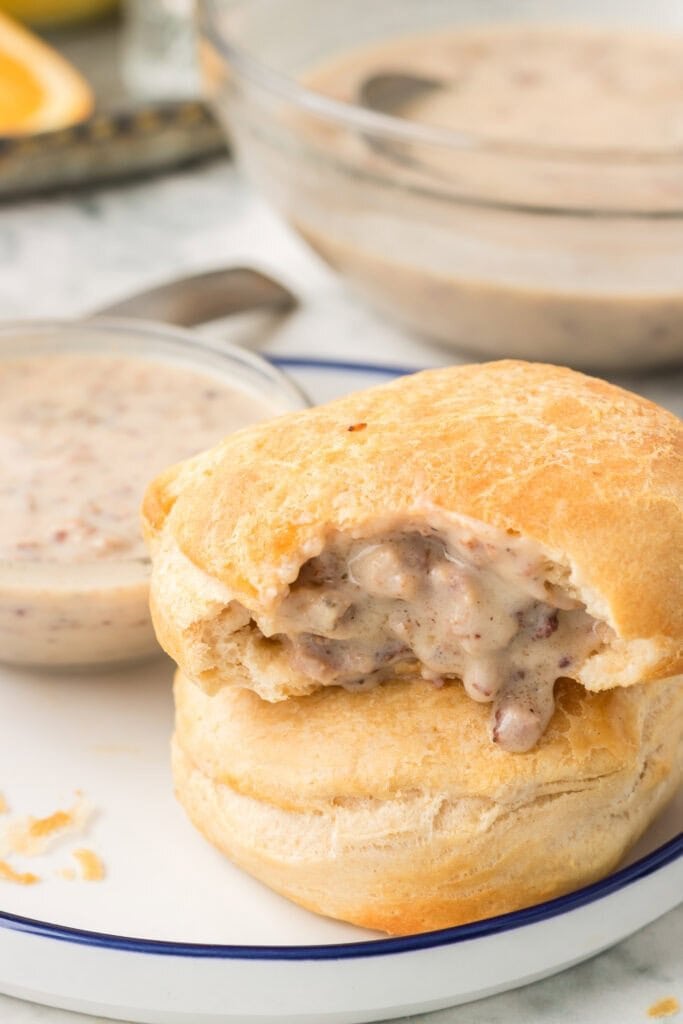 A biscuit with creamy sausage gravy filling, sitting on a white plate. The top biscuit has a bite taken out, showing the gravy inside. A bowl of extra gravy is in the background.