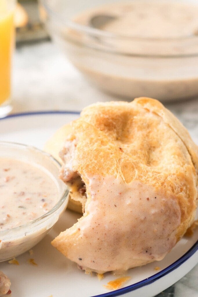 An Air Fryer Sausage Gravy Stuffed Biscuit with a bite taken out sits on a white plate. A small bowl of extra gravy is beside it, and a glass of orange juice is in the background.