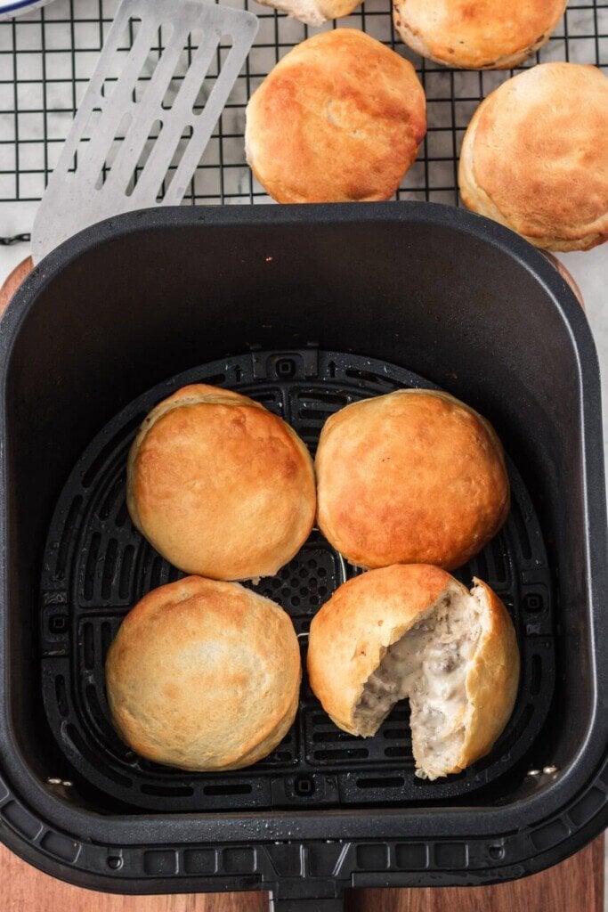 Four Air Fryer Sausage Gravy Stuffed Biscuits are nestled in an air fryer basket, with one revealing its creamy, savory filling inside. More stuffed biscuits and a spatula rest on a cooling rack in the background, ready to enjoy.