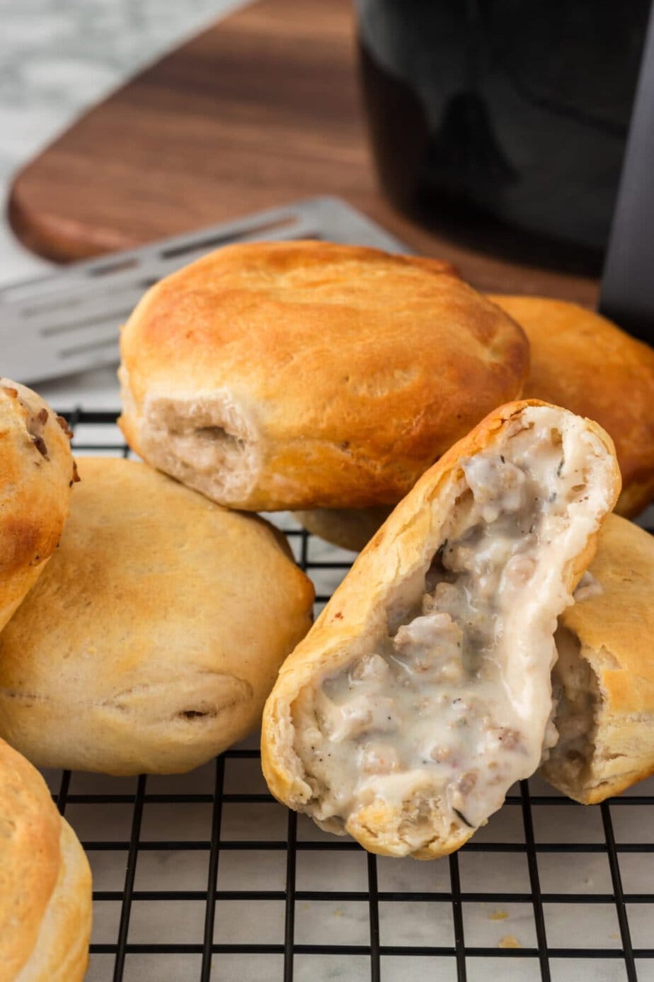 Air Fryer Sausage Gravy Stuffed Biscuits are golden-brown biscuit rolls filled with creamy sausage gravy, resting on a cooling rack; one roll is cut open, revealing the savory filling inside.