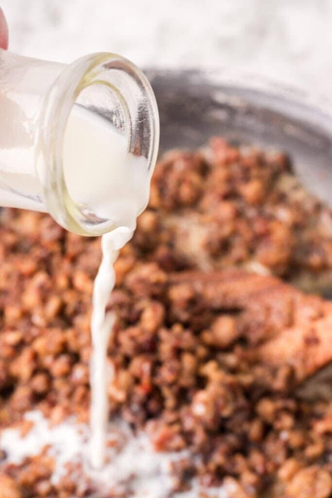A close-up of milk being poured from a small glass bottle onto a mixture of cooked ground sausage in a pan.