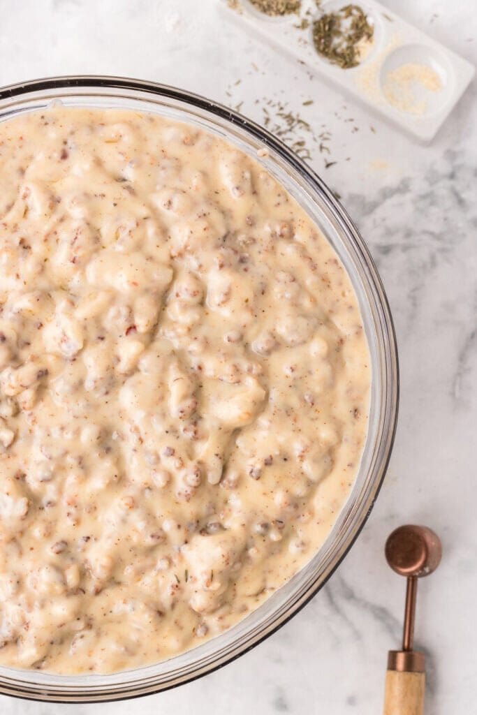 A bowl filled with creamy sausage gravy sits on a marble surface next to a wooden measuring spoon and a small dish of dried herbs and spices.