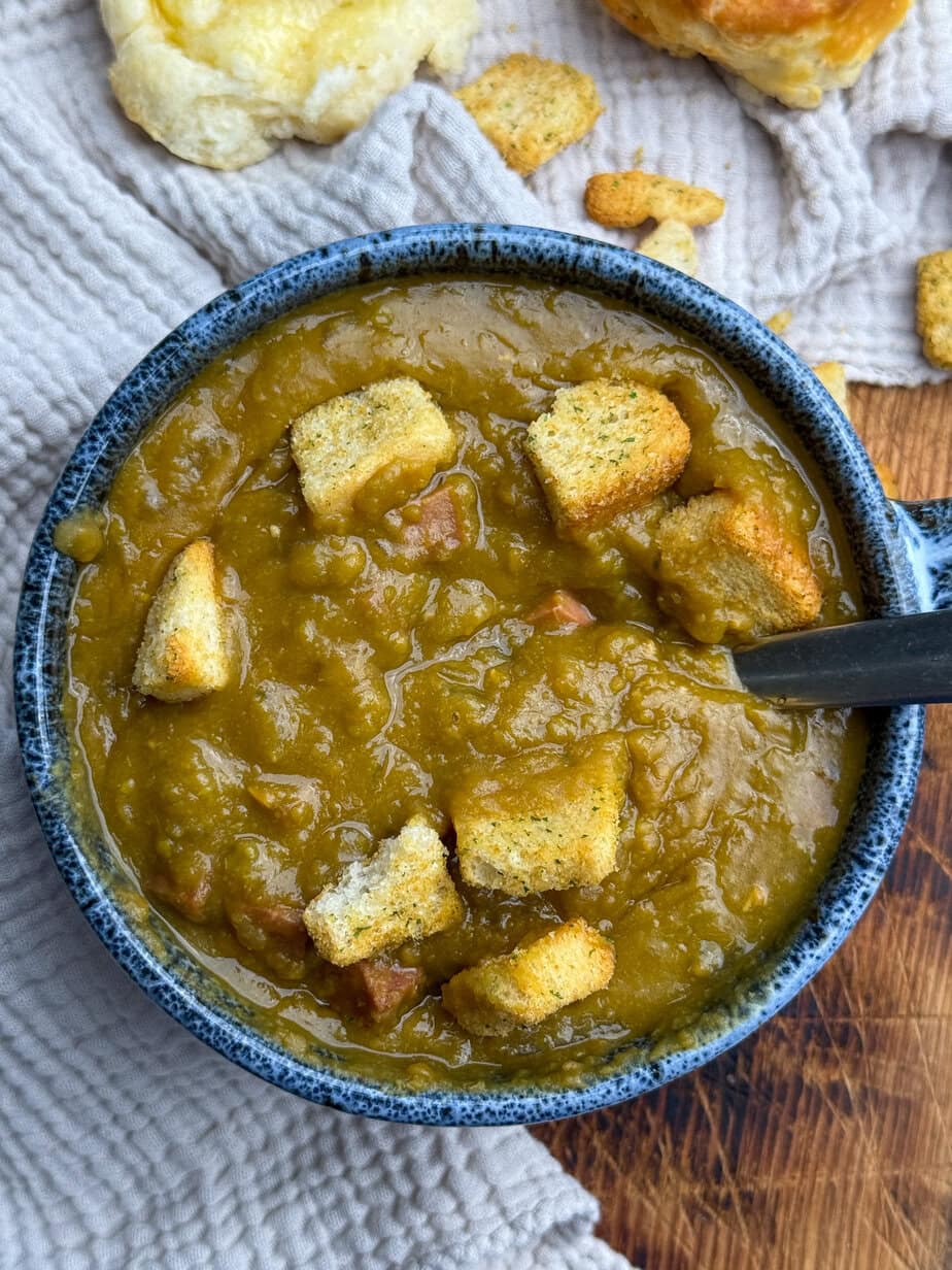 A blue bowl of thick split pea soup with croutons on top sits on a textured beige cloth, with a spoon in the bowl and pieces of bread nearby.