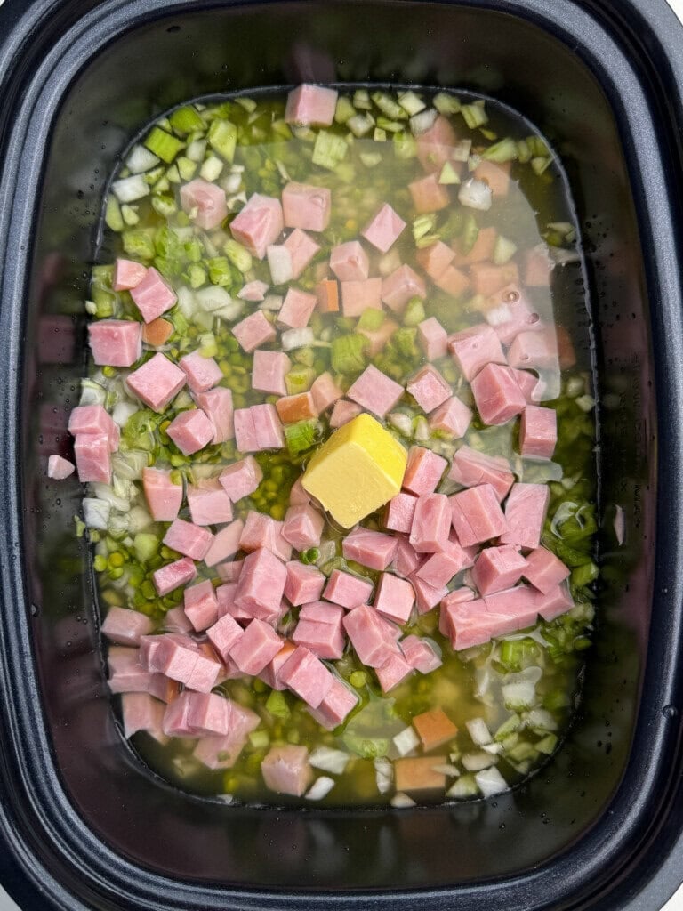 Cubes of ham, chopped celery, onion, carrots, and a pat of butter are in a slow cooker with broth, ready to cook.