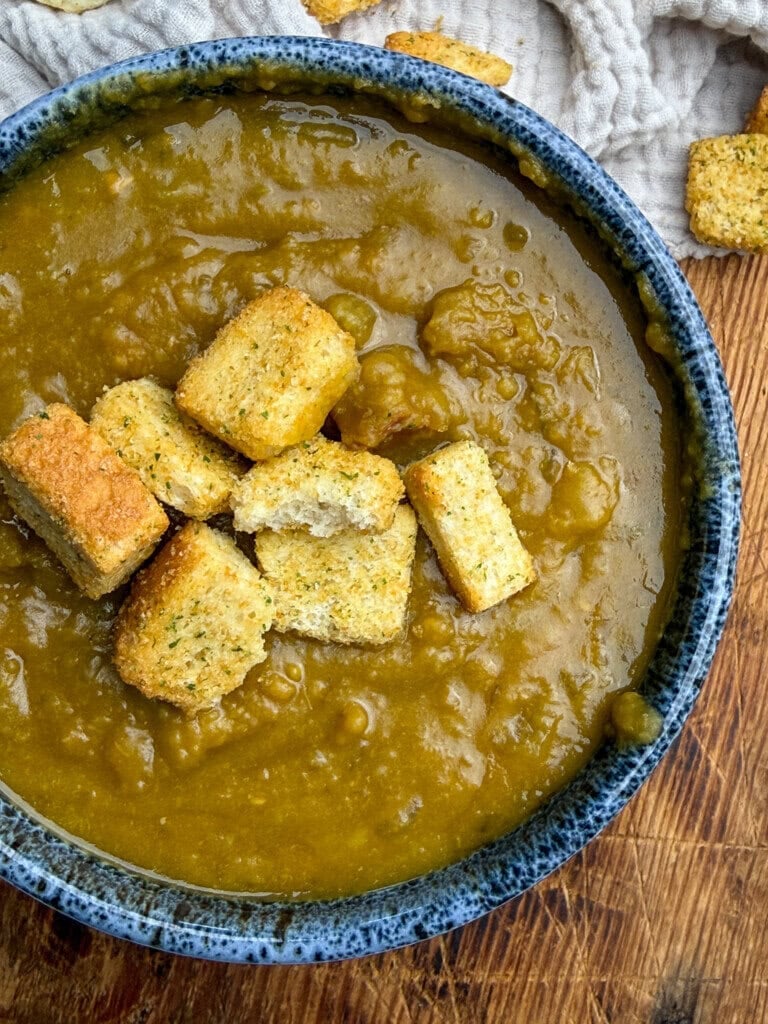 A blue bowl of thick, brownish-green split pea soup topped with several golden croutons sits on a wooden surface, with a light-colored textured cloth and more croutons in the background.