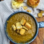 A blue bowl of thick soup topped with croutons sits on a wooden board next to two golden biscuits, one split open with butter, all on a textured beige cloth.