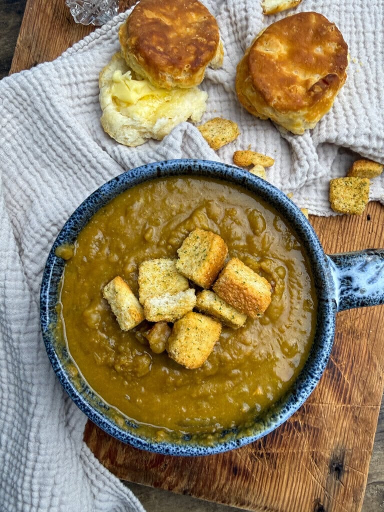 A blue bowl of thick soup topped with croutons sits on a wooden board next to two golden biscuits, one split open with butter, all on a textured beige cloth.