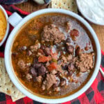 A bowl of chili with ground beef, black beans, and tomatoes sits on a red plaid cloth, surrounded by tortilla chips, shredded cheese, and a bowl of sour cream.