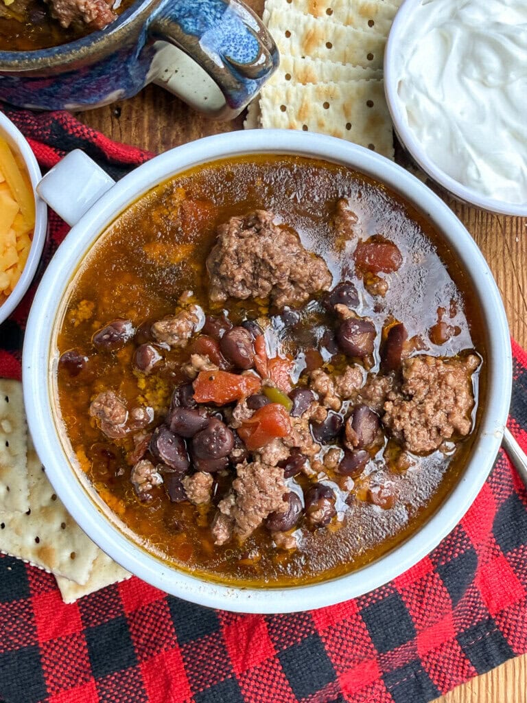 A bowl of chili with ground beef, black beans, and tomatoes sits on a red plaid cloth, surrounded by saltine crackers, shredded cheese, and a bowl of sour cream.