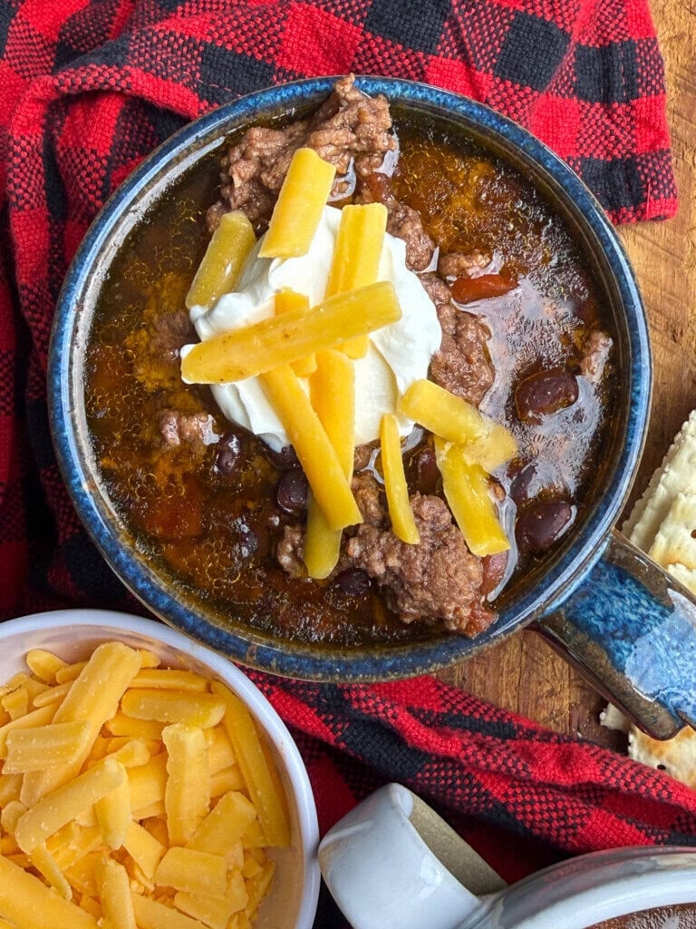 A bowl of chili topped with shredded cheese and sour cream sits on a wooden surface next to a red plaid cloth, a bowl of cheddar cheese, and some crackers.