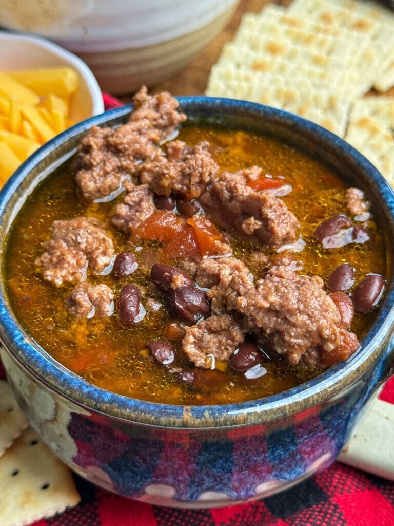 A bowl of chili with ground beef, beans, and tomatoes sits next to a plate of crackers and shredded cheddar cheese.