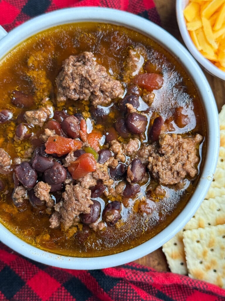 A bowl of chili with ground beef, black beans, and tomatoes sits on a red plaid cloth, with shredded cheese and saltine crackers on the side.