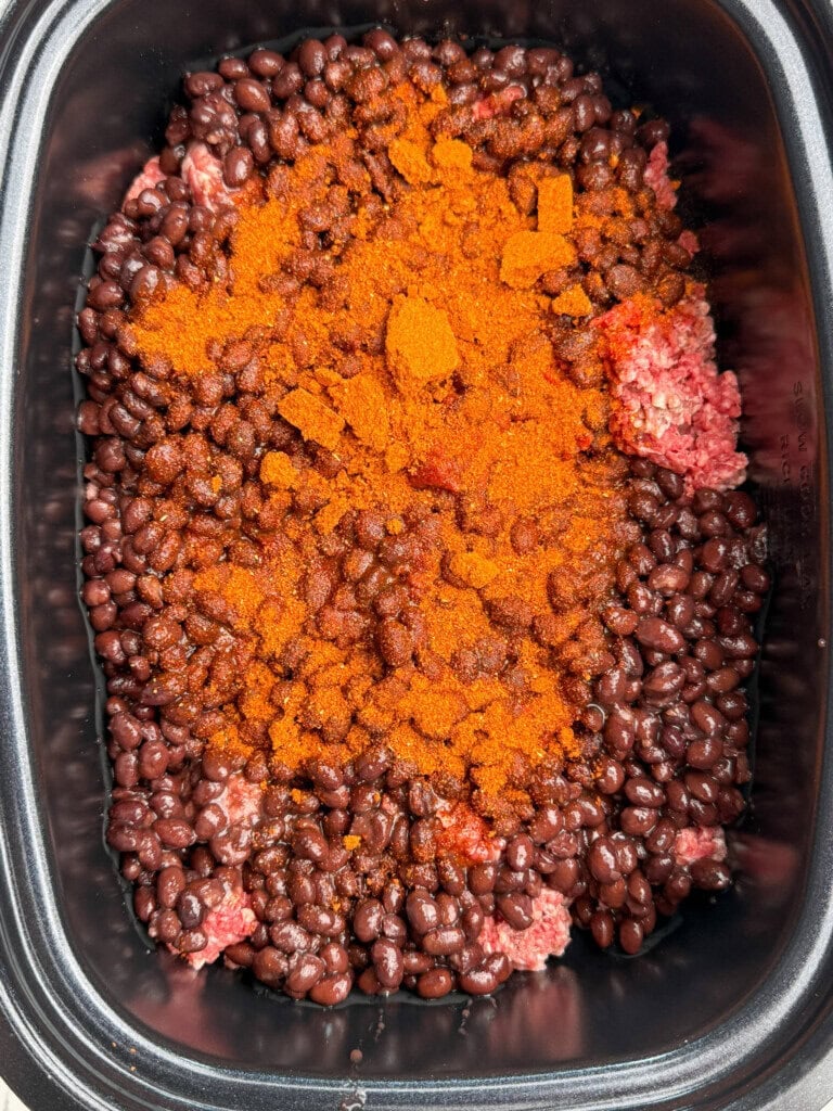 Ground beef, black beans, and a generous amount of chili powder seasoning in a slow cooker, ready to be mixed and cooked.