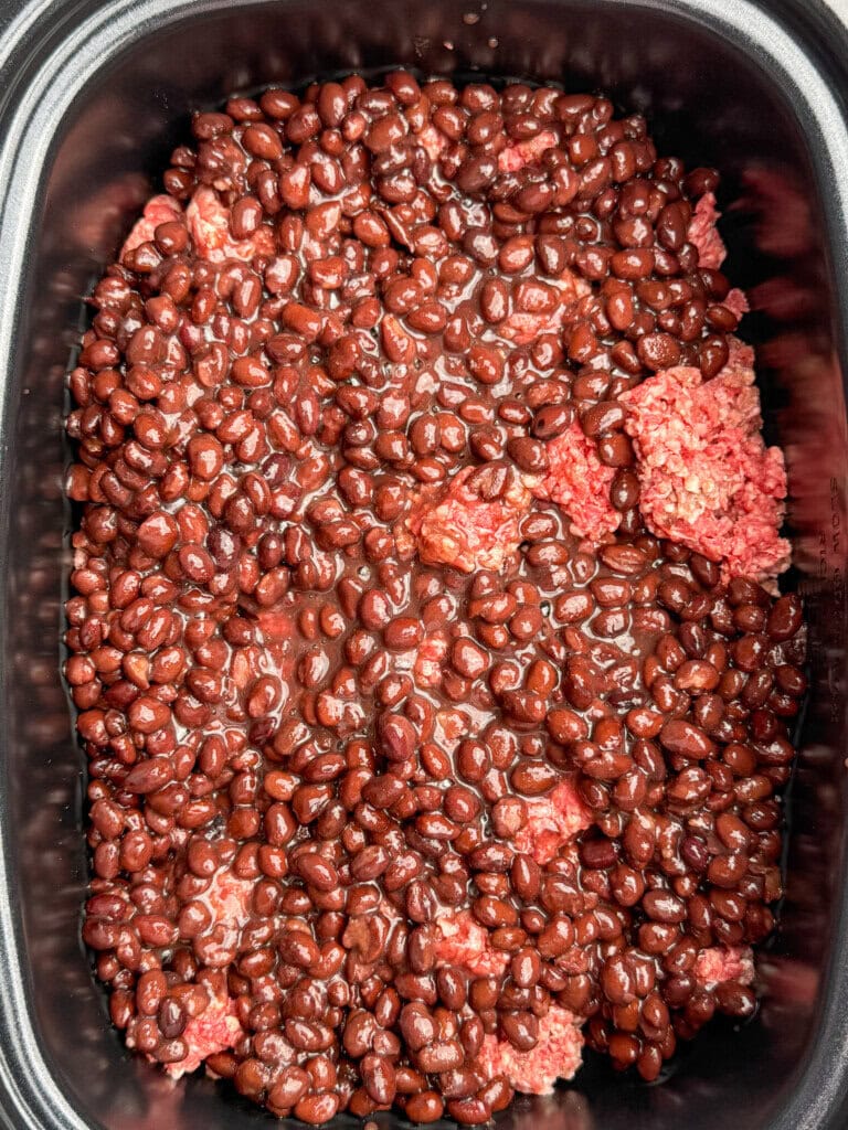 Raw ground beef topped with a layer of canned black beans in a slow cooker, ready to be cooked.
