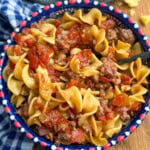 A colorful bowl filled with egg noodles, ground beef, and diced tomatoes sits on a wooden table beside a blue-checkered cloth. A spoon is in the bowl, and cornbread is visible on a plate in the background.