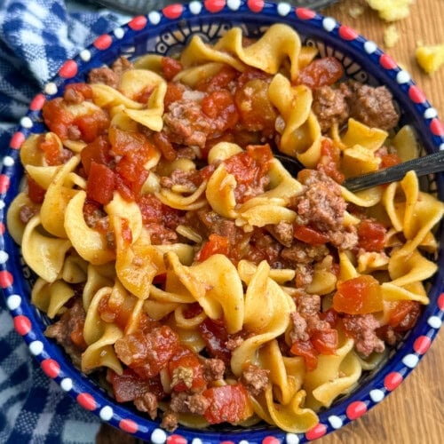 A colorful bowl filled with egg noodles, ground beef, and diced tomatoes sits on a wooden table beside a blue-checkered cloth. A spoon is in the bowl, and cornbread is visible on a plate in the background.