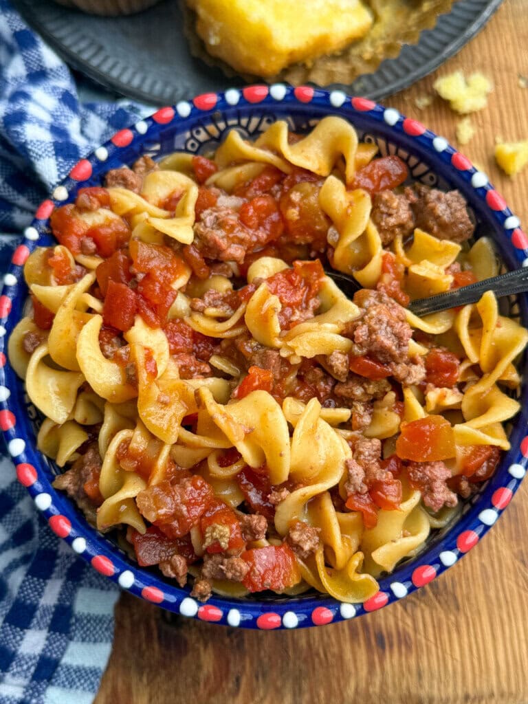 A colorful bowl filled with egg noodles, ground beef, and diced tomatoes sits on a wooden table beside a blue-checkered cloth. A spoon is in the bowl, and cornbread is visible on a plate in the background.