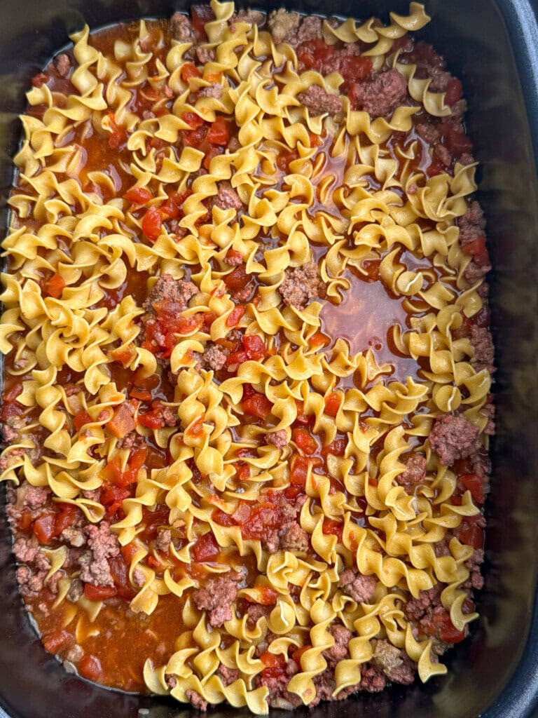A crock pot dish filled with uncooked egg noodles, ground beef, diced tomatoes, and sauce, ready to be cooked.