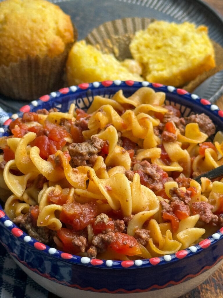 A bowl of beef and tomato noodle casserole sits in the foreground, with two pieces of golden cornbread on a plate in the background. A spoon is placed inside the bowl.