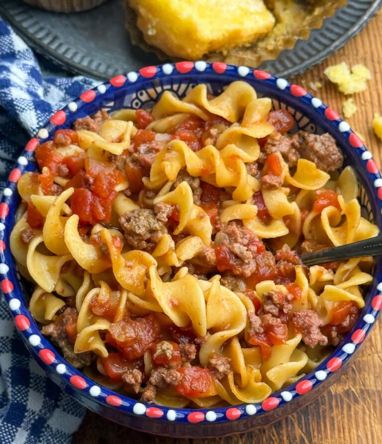 A bowl of pasta with ground beef and diced tomatoes sits on a table with a blue checkered napkin and cornbread in the background. A black fork rests inside the bowl.