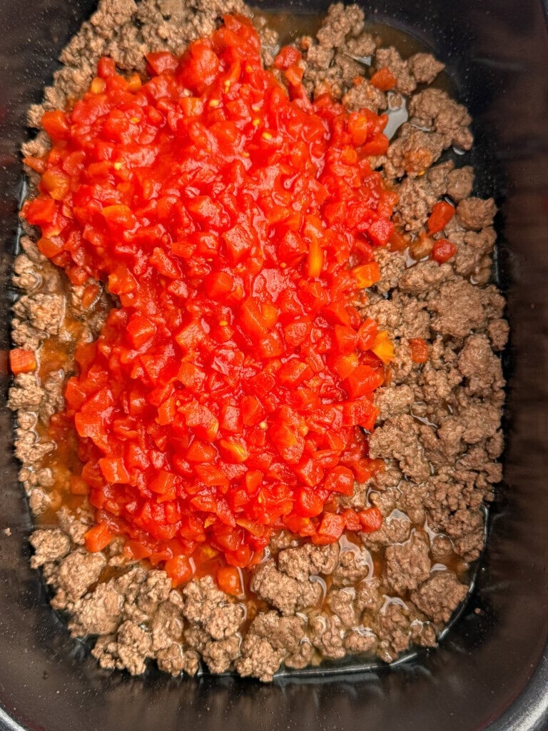 Cooked ground beef topped with a generous amount of finely diced tomatoes in a slow cooker.