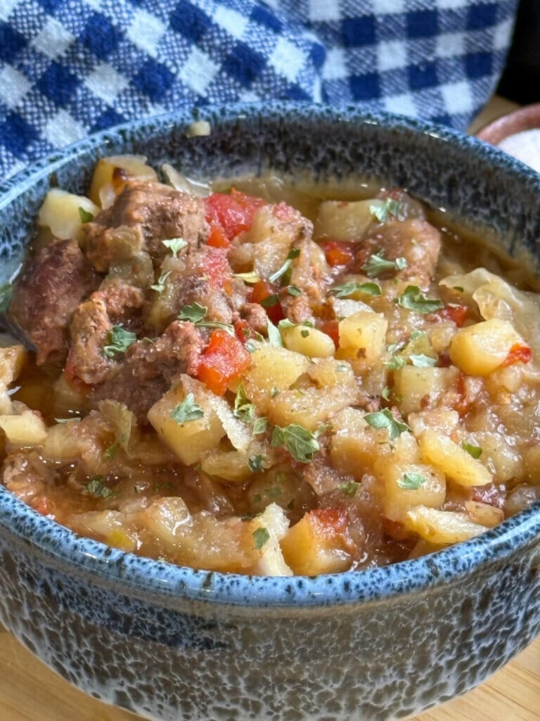 A blue bowl filled with chunky beef stew containing potatoes, carrots, and herbs sits on a wooden surface next to a blue and white checkered cloth.