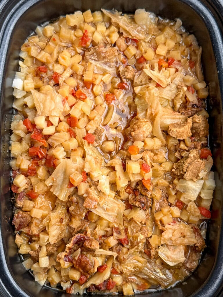 A close-up of a dish with diced potatoes, cabbage, small pieces of meat, and tomatoes in a savory sauce, all cooked together in a black crock pot.