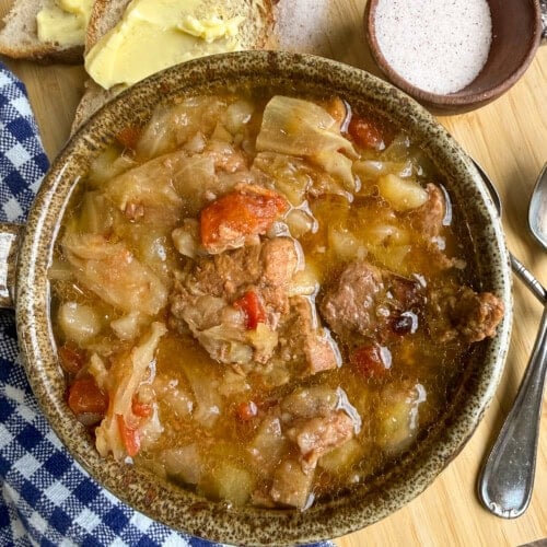 A bowl of stew with meat, cabbage, and tomatoes sits on a wooden board, next to buttered bread, a small bowl of salt, a spoon, and a blue checkered napkin.