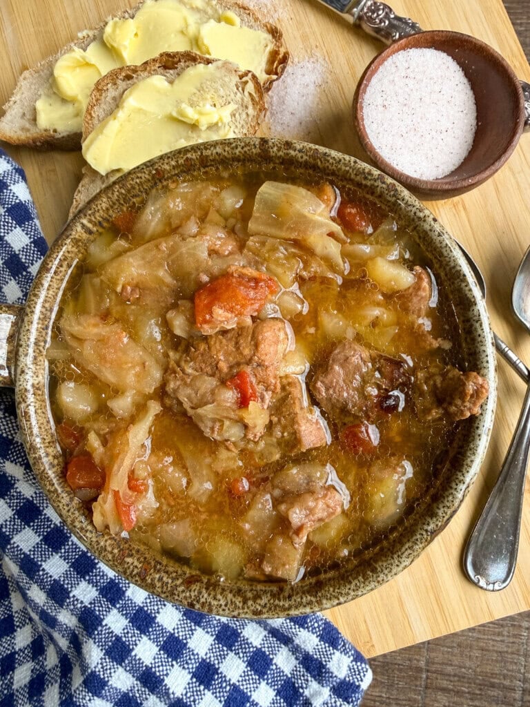 A bowl of stew with meat, cabbage, and tomatoes sits on a wooden board, next to buttered bread, a small bowl of salt, a spoon, and a blue checkered napkin.