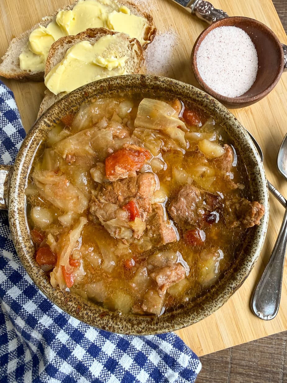 A bowl of stew with meat, cabbage, and tomatoes sits on a wooden board, next to buttered bread, a small bowl of salt, a spoon, and a blue checkered napkin.