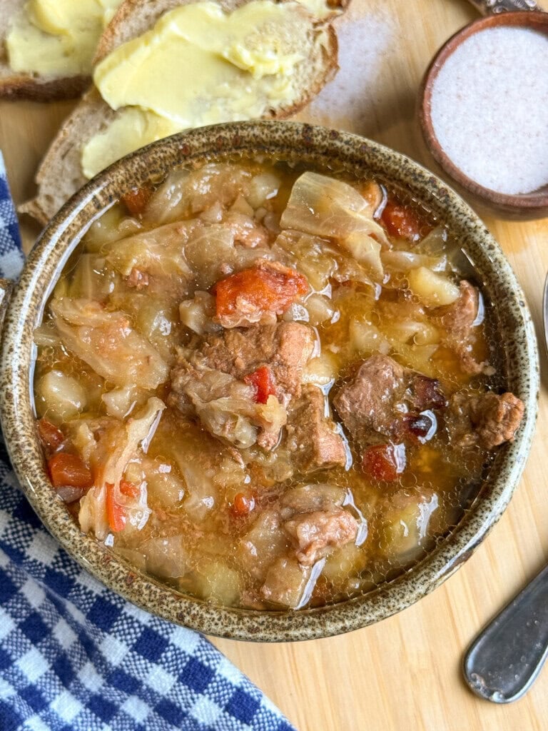 A bowl of hearty stew with chunks of meat, cabbage, and vegetables, sits next to buttered bread and a small dish of salt on a wooden surface with a blue checkered cloth.