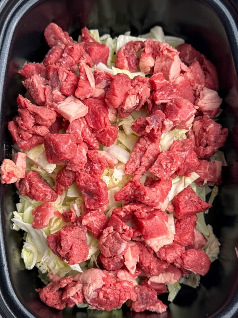 Chunks of raw beef are spread over a bed of chopped cabbage inside a black slow cooker or pot, ready for cooking.