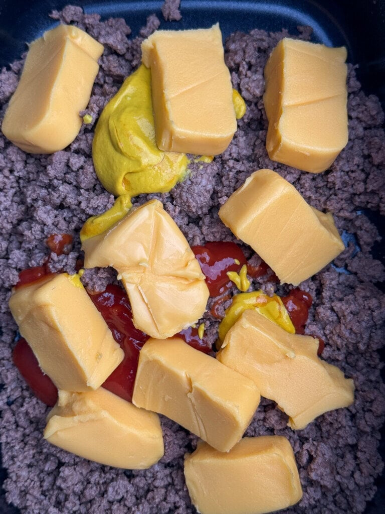 Cooked ground beef in a pan topped with slices of processed cheese, yellow mustard, and ketchup, about to be mixed together.