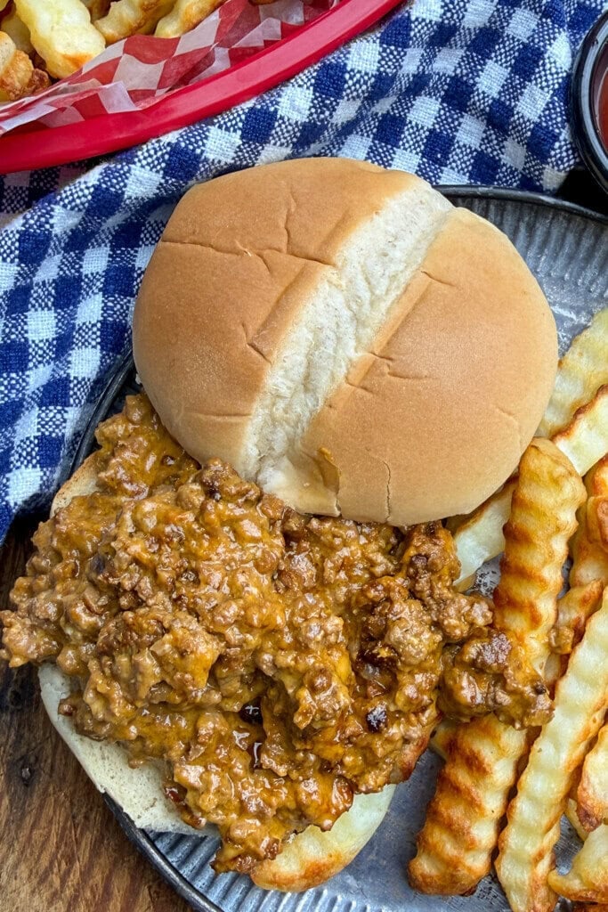 A sloppy joe sandwich with seasoned ground beef on a bun, served with crinkle-cut fries on a metal plate. A blue and white checkered cloth and part of a basket of fries are in the background.