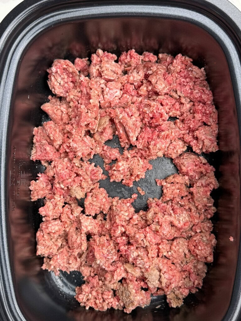 Raw ground beef spread unevenly in the bottom of a black slow cooker, with some portions starting to brown while most remain pink and uncooked.