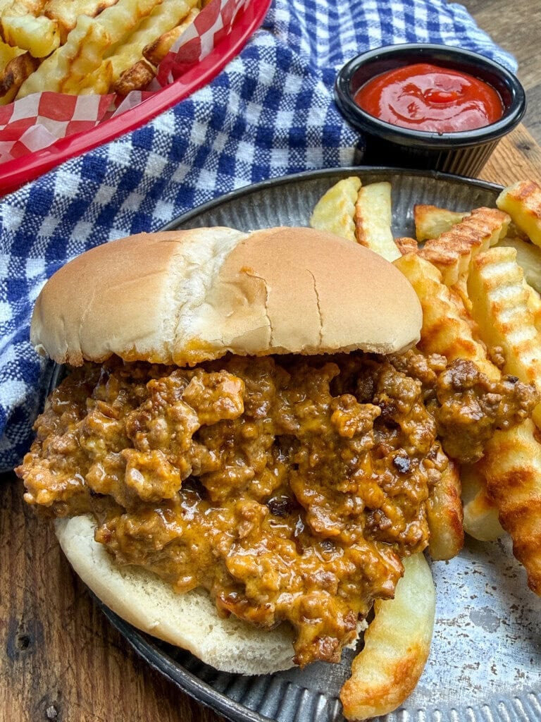 A plate with a sloppy joe sandwich and crinkle-cut fries sits on a metal tray, with a small cup of ketchup and a basket of fries nearby, on a blue checkered cloth.