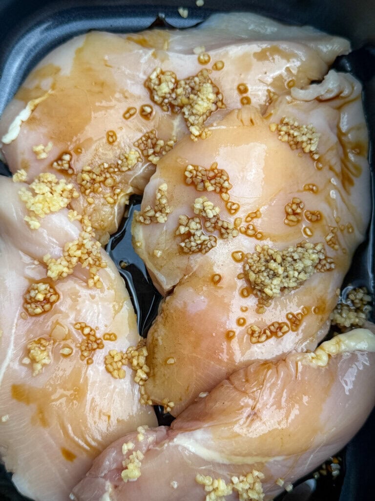 Raw chicken breasts in a black slow cooker, topped with minced garlic and drizzled with a brown marinade or sauce.