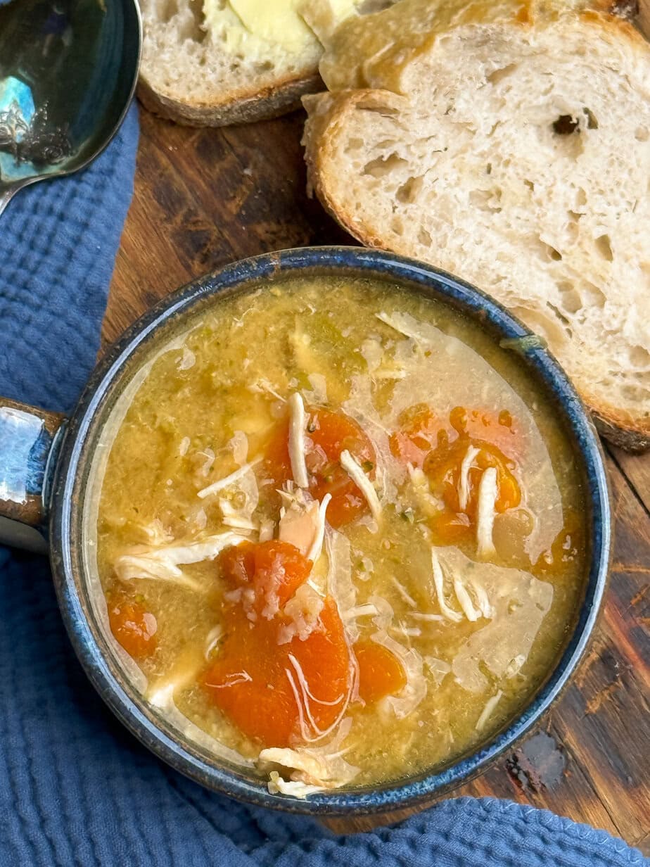 A bowl of chicken soup with shredded chicken and carrots, served on a blue cloth next to slices of buttered bread on a wooden surface.