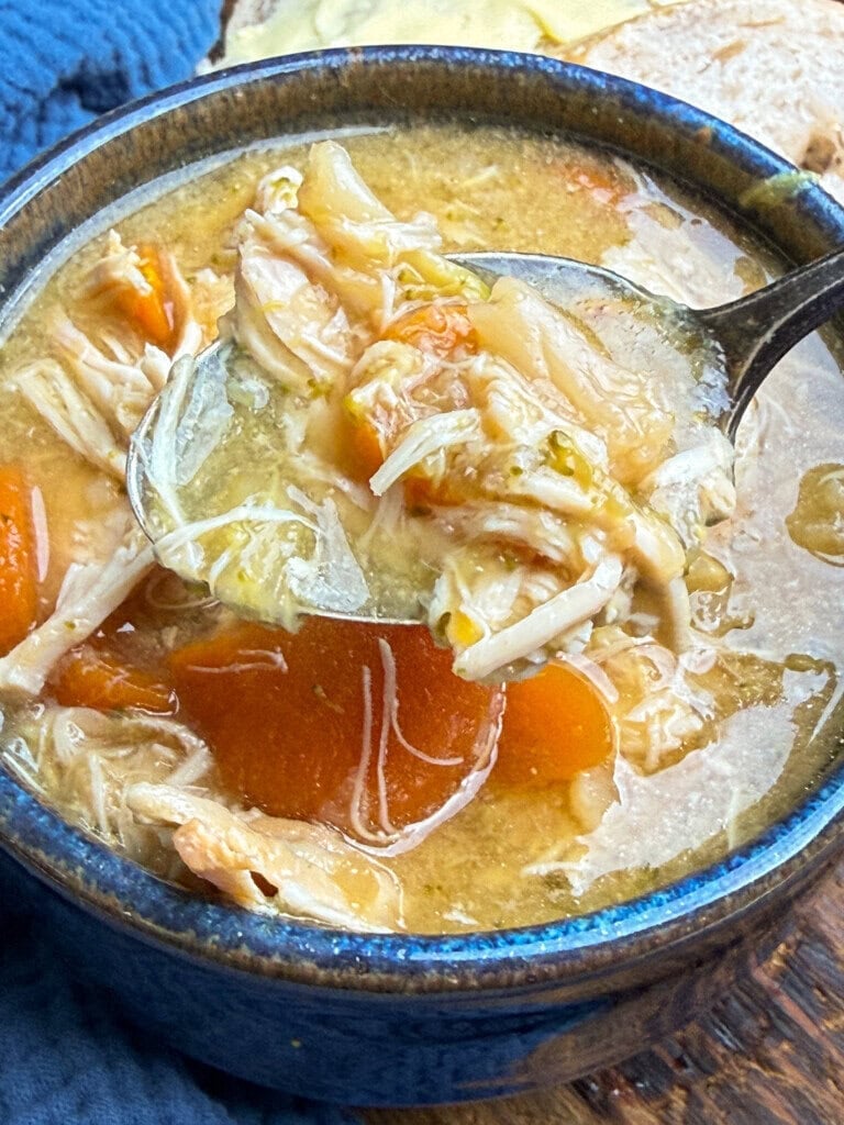 A close-up of a bowl of chicken soup with shredded chicken, carrots, and broth. A spoon is lifting a portion from the bowl.