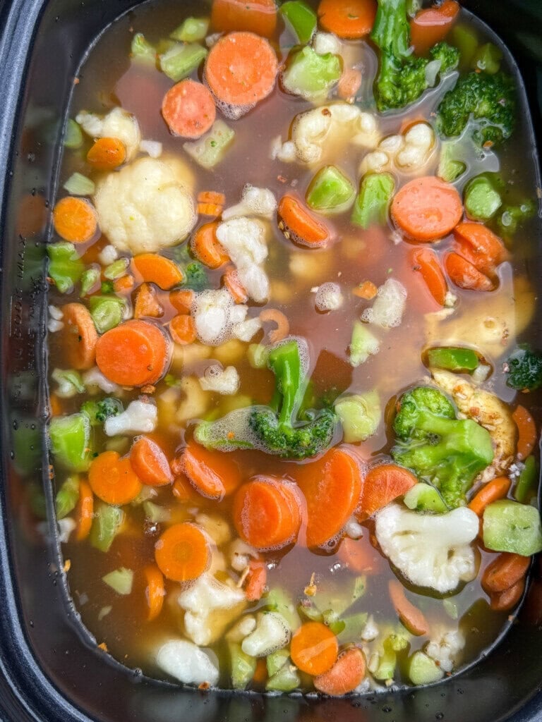Chopped carrots, broccoli, and cauliflower pieces floating in a brownish broth inside a slow cooker or pot, ready to be cooked as a vegetable soup or stew.