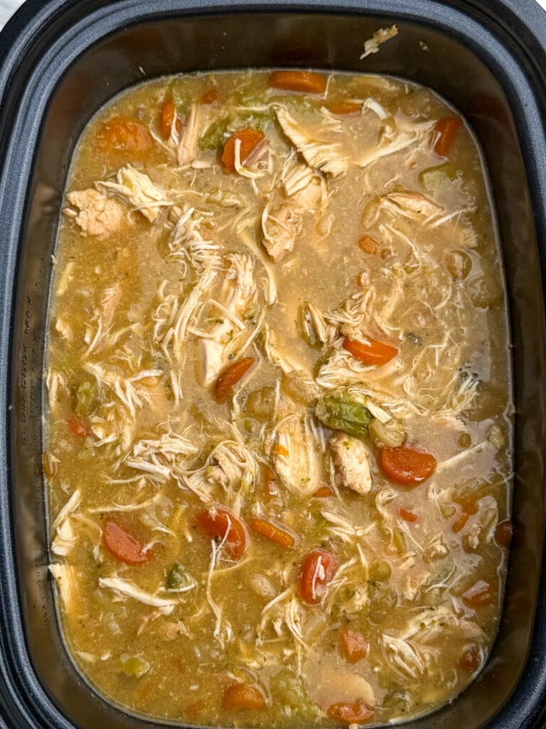 Shredded chicken, sliced carrots, and celery in a thick, golden broth inside a black slow cooker, resembling homemade chicken stew or soup.